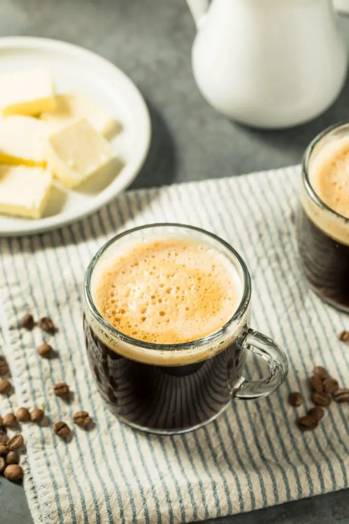 Two glass mugs of frothy butter coffee on a striped cloth, with coffee beans scattered around and a plate of butter in the background.