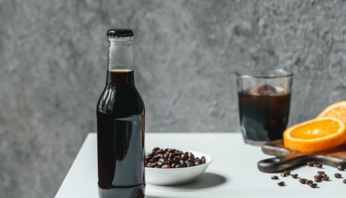 A bottle of cold brew, a bowl of coffee beans, a glass of the beverage, and sliced oranges are arranged on a table with a gray textured background..