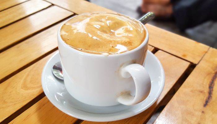 A white ceramic cup filled with Vietnamese egg coffee on a matching saucer with a metal spoon, placed on a wooden table.