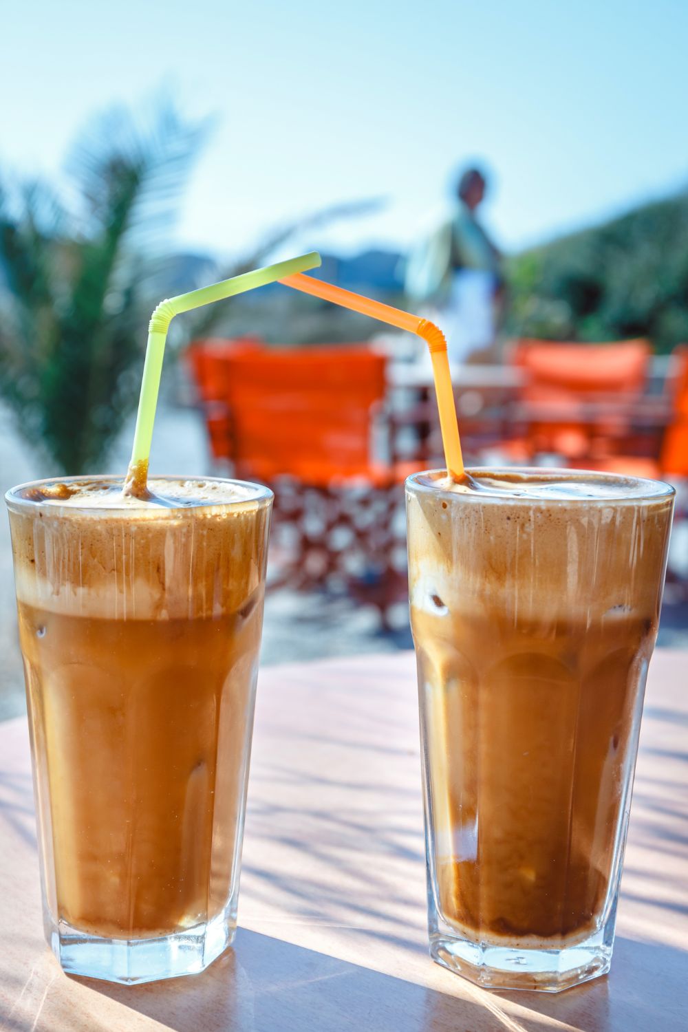 Two glasses of iced frappe with straws on an outdoor table, with a blurry background of a person walking near tables.