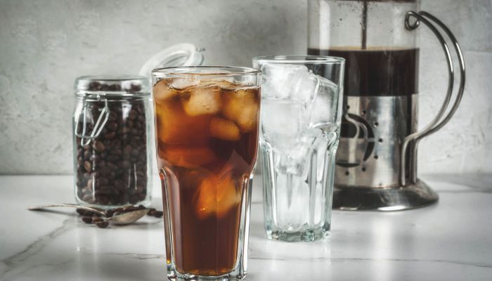 A glass of iced coffee sits next to a French press and a jar of coffee beans, with a cup of ice on a marble surface.