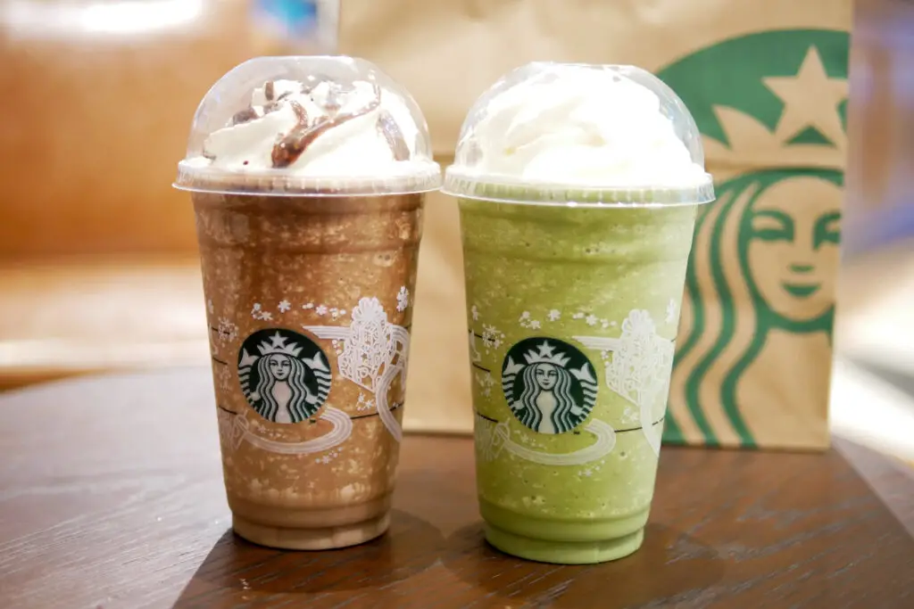Two Starbucks drinks, a mocha frappuccino and a matcha frappuccino, on a wooden table with a Starbucks logo in the background.