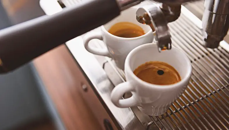 Two white ceramic cups of espresso being pulled from machine.