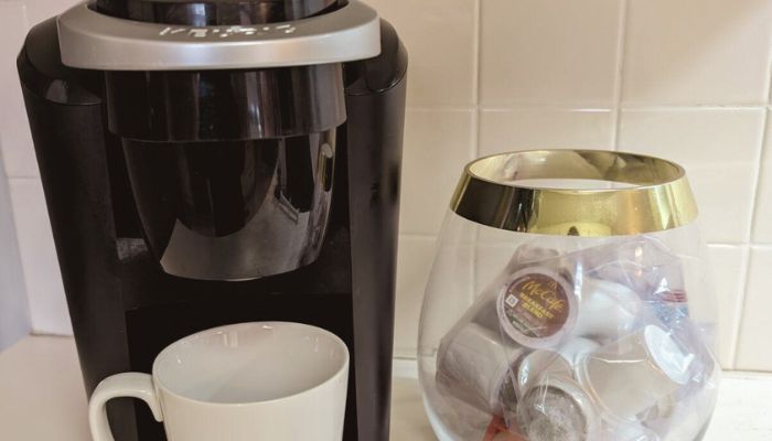 A black single-serve Keurig coffee maker stands beside a glass bowl filled with coffee pods. A white ceramic mug waits under the machine, ready for your next espresso.
