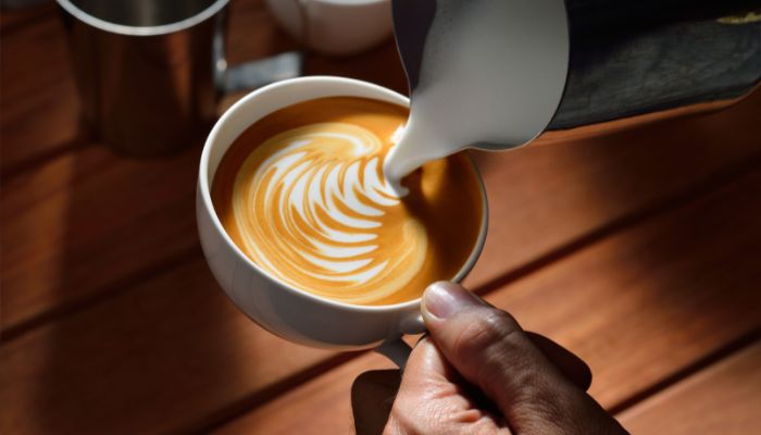 A hand pouring steamed milk into a cup of breve coffee, creating a latte art pattern on the surface.